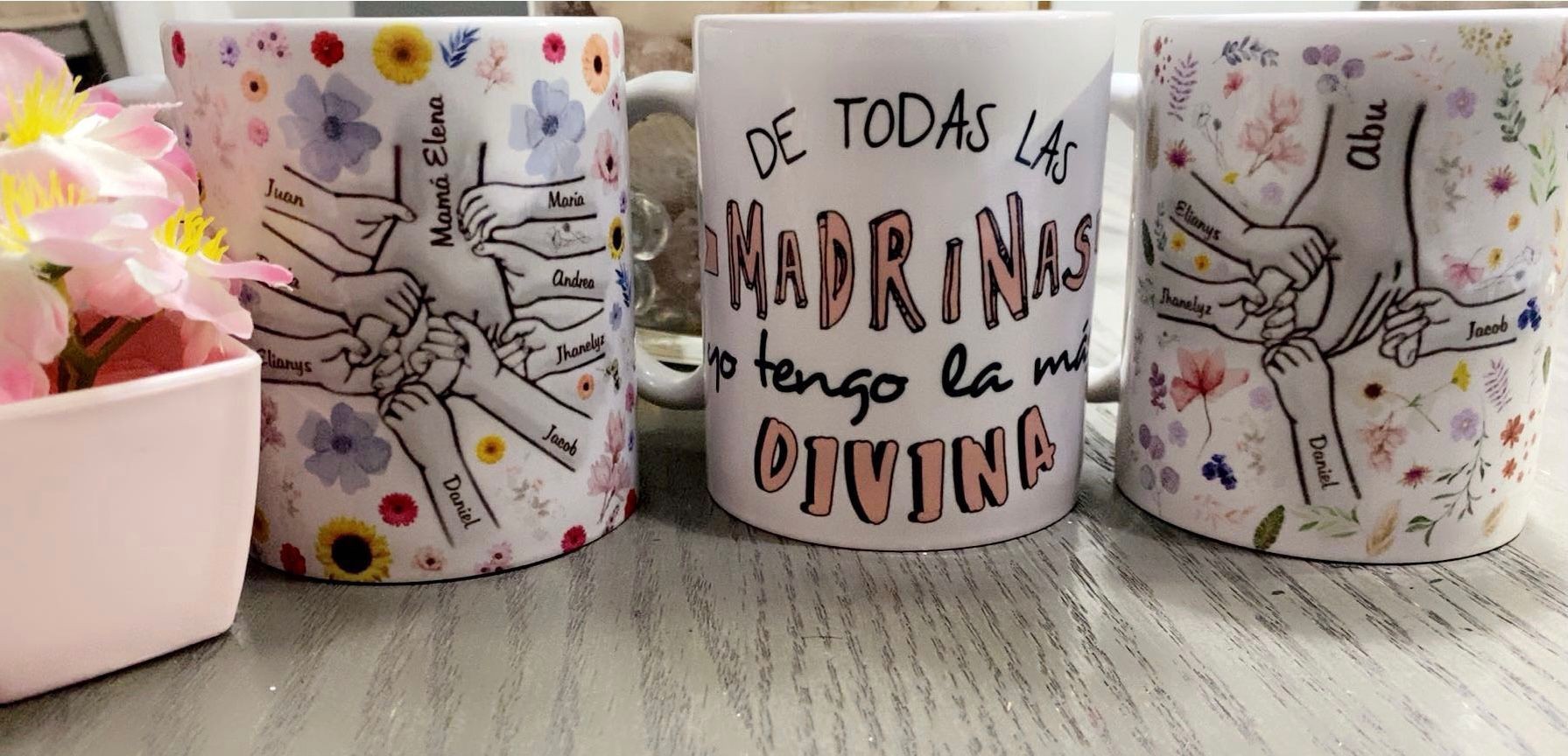 Three personalized mugs with illustrations of family members holding hands and Spanish text surrounded by pink flowers.